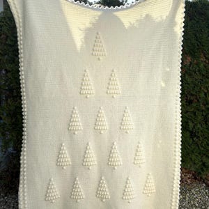 May include: A white crocheted blanket with a repeating pattern of Christmas trees made with raised stitches. The blanket has a scalloped edge.