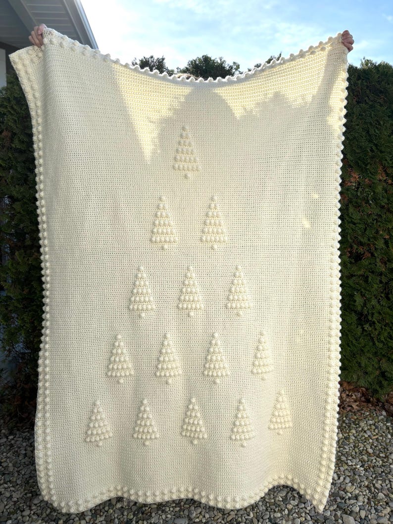 May include: A white crocheted blanket with a repeating pattern of Christmas trees made with raised stitches. The blanket has a scalloped edge.