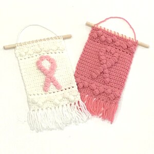 Puede incluir: Dos tapices de pared de ganchillo con hilo rosa y blanco. El tapiz blanco presenta un dise&ntilde;o de cinta rosa. Ambos tapices tienen flecos en la parte inferior.