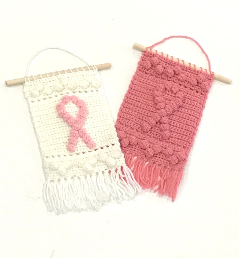 Puede incluir: Dos tapices de pared de ganchillo con hilo rosa y blanco. El tapiz blanco presenta un dise&ntilde;o de cinta rosa. Ambos tapices tienen flecos en la parte inferior.