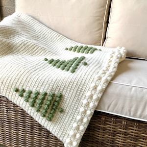 May include: A white crocheted throw blanket with a green geometric design. The blanket has a white crocheted border with small white pom-poms.