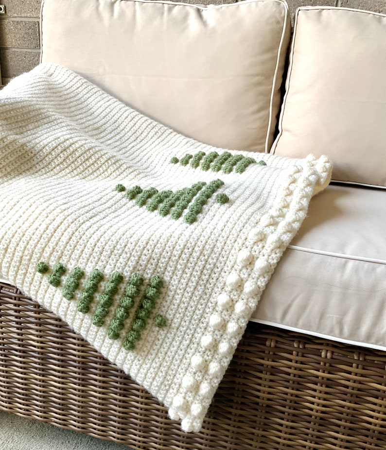 May include: A white crocheted throw blanket with a green geometric design. The blanket has a white crocheted border with small white pom-poms.