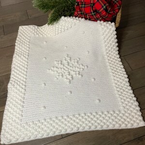 May include: White crocheted baby blanket with a snowflake design in the center. The blanket has a textured border with raised bobbles.
