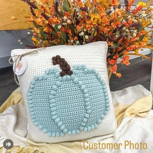 May include: A square white crochet pillow featuring a large, textured light blue pumpkin appliqu&eacute; with a brown stem. A small round tag with a graphic illustration is attached to the top left corner with a thin brown twine string.