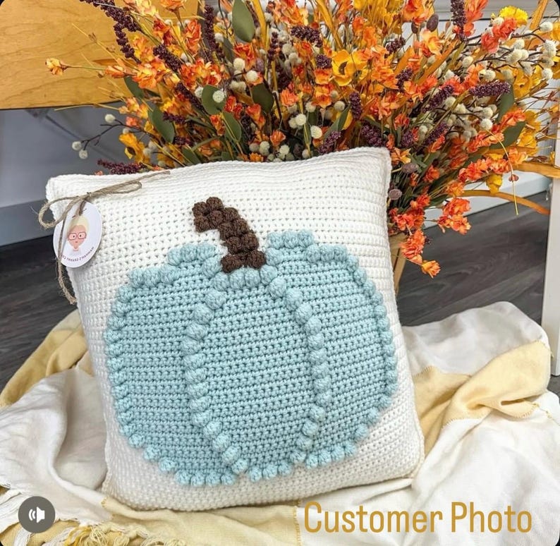 May include: A square white crochet pillow featuring a large, textured light blue pumpkin appliqu&eacute; with a brown stem. A small round tag with a graphic illustration is attached to the top left corner with a thin brown twine string.
