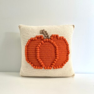 May include: A square crochet pillow in off-white featuring a centered, textured orange pumpkin design. The pumpkin is outlined with small, raised orange bobble stitches and includes a small, brown, textured stem at the top center.