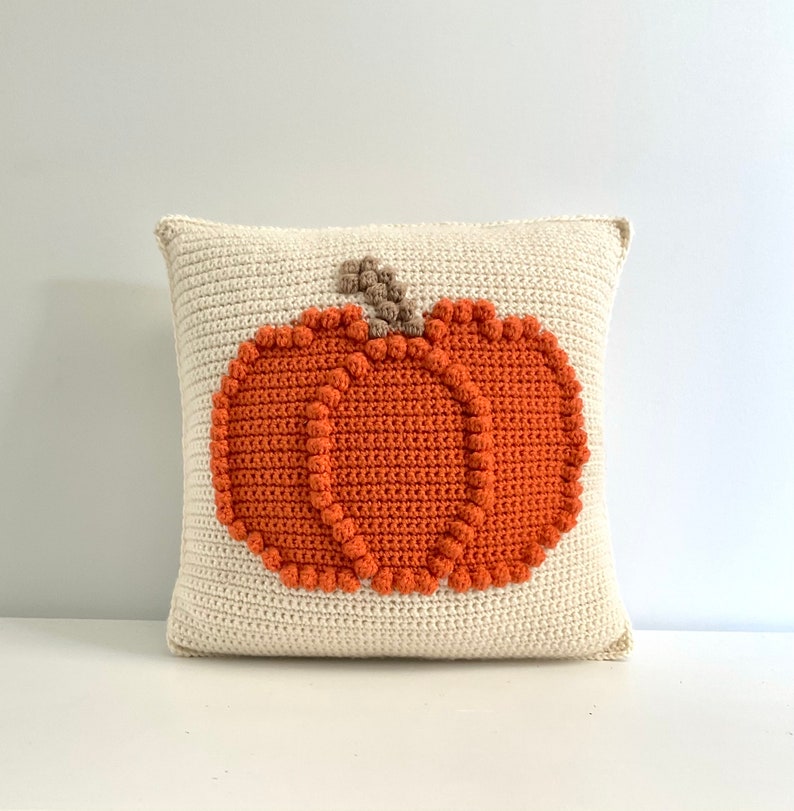 May include: A square crochet pillow in off-white featuring a centered, textured orange pumpkin design. The pumpkin is outlined with small, raised orange bobble stitches and includes a small, brown, textured stem at the top center.