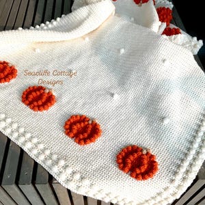 May include: A cream-colored, crocheted blanket with orange pumpkin appliqu&eacute;s. The blanket features a textured stitch pattern and a decorative border with bobble stitches. The text "Seacliff Cottage Designs" is visible on the blanket.