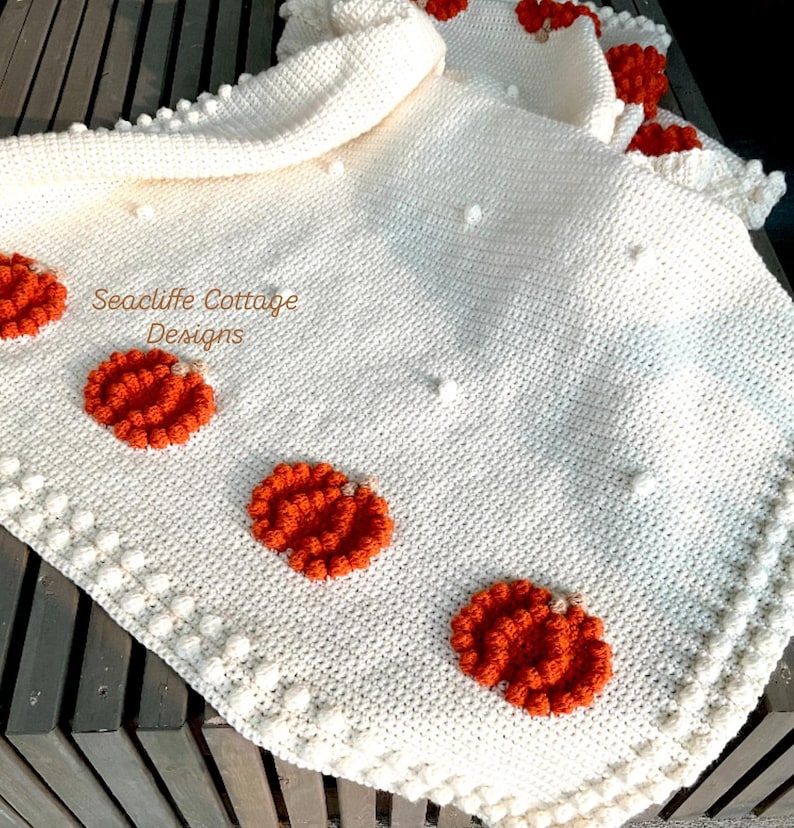 May include: A cream-colored, crocheted blanket with orange pumpkin appliqu&eacute;s. The blanket features a textured stitch pattern and a decorative border with bobble stitches. The text "Seacliff Cottage Designs" is visible on the blanket.