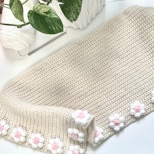 May include: A crocheted beige blanket with a white and pink flower border.