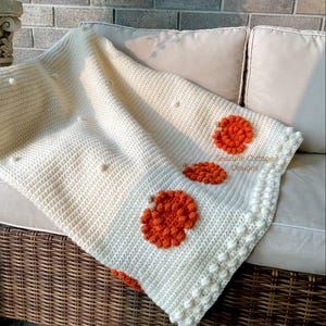 May include: Cream-colored crochet blanket with three orange floral designs. The blanket has a textured stitch pattern and a decorative border. The text "Seacliffe Cottage Designs" is visible on the blanket.