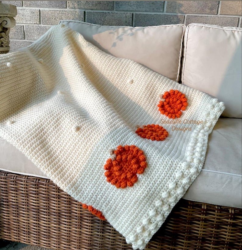 May include: Cream-colored crochet blanket with three orange floral designs. The blanket has a textured stitch pattern and a decorative border. The text "Seacliffe Cottage Designs" is visible on the blanket.