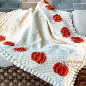 May include: Cream-colored crochet blanket with orange floral accents. The blanket features a textured stitch pattern, a scalloped edge, and raised bobble details. The blanket is draped over a wicker basket and a white couch.