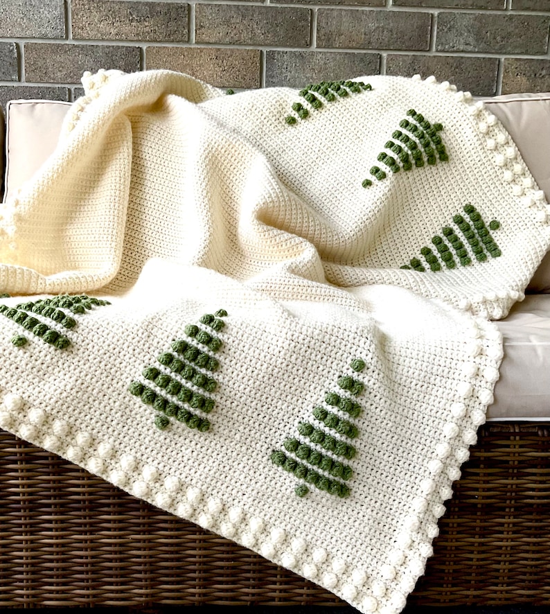 May include: A white crocheted throw blanket with a green embroidered design of Christmas trees. The blanket has a white pom-pom trim.
