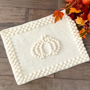 May include: Cream-colored, rectangular crochet blanket with a pumpkin design in the center. The blanket features a textured border and a raised pumpkin motif, perfect for fall decor. The blanket is made of soft yarn.