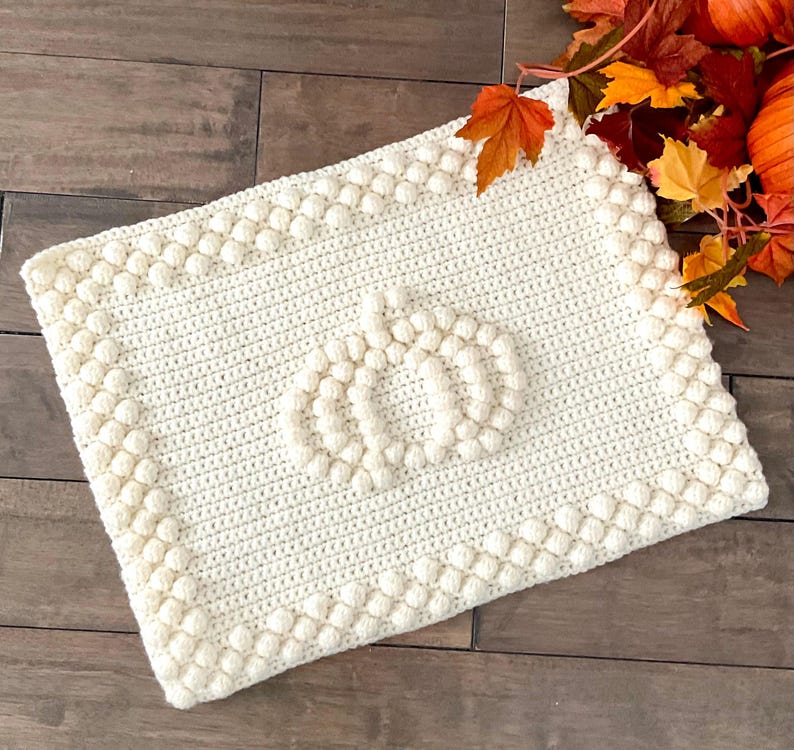 May include: Cream-colored, rectangular crochet blanket with a pumpkin design in the center. The blanket features a textured border and a raised pumpkin motif, perfect for fall decor. The blanket is made of soft yarn.