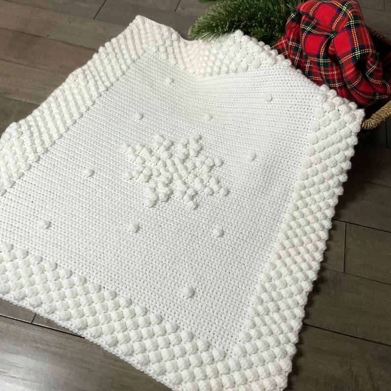 May include: White crocheted blanket with a snowflake design in the center. The blanket has a textured border with raised bobbles.