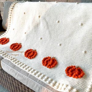 May include: Cream-colored crochet blanket with orange pumpkin appliqu&eacute;s. The blanket features a textured design with bobble stitch accents and is draped over a gray sofa. The blanket has the text "Seacliff Cottage Designs".