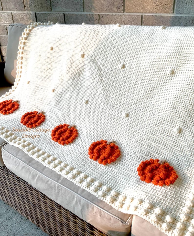 May include: Cream-colored crochet blanket with orange pumpkin appliqu&eacute;s. The blanket features a textured design with bobble stitch accents and is draped over a gray sofa. The blanket has the text "Seacliff Cottage Designs".