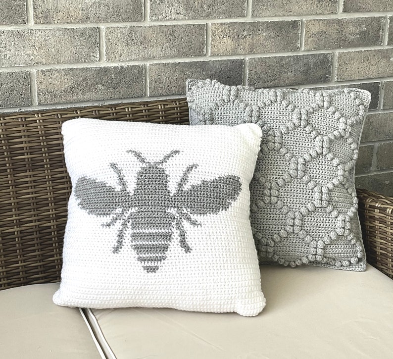 May include: Two decorative throw pillows, one white with a gray crocheted bee design and one gray with a crocheted geometric pattern. The pillows are on a wicker couch.