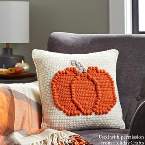 May include: A square, cream-colored crochet pillow featuring a textured, raised orange pumpkin design in the center with a small grey stem. The pumpkin is constructed with a bobble stitch pattern, creating a distinct, three-dimensional appearance.