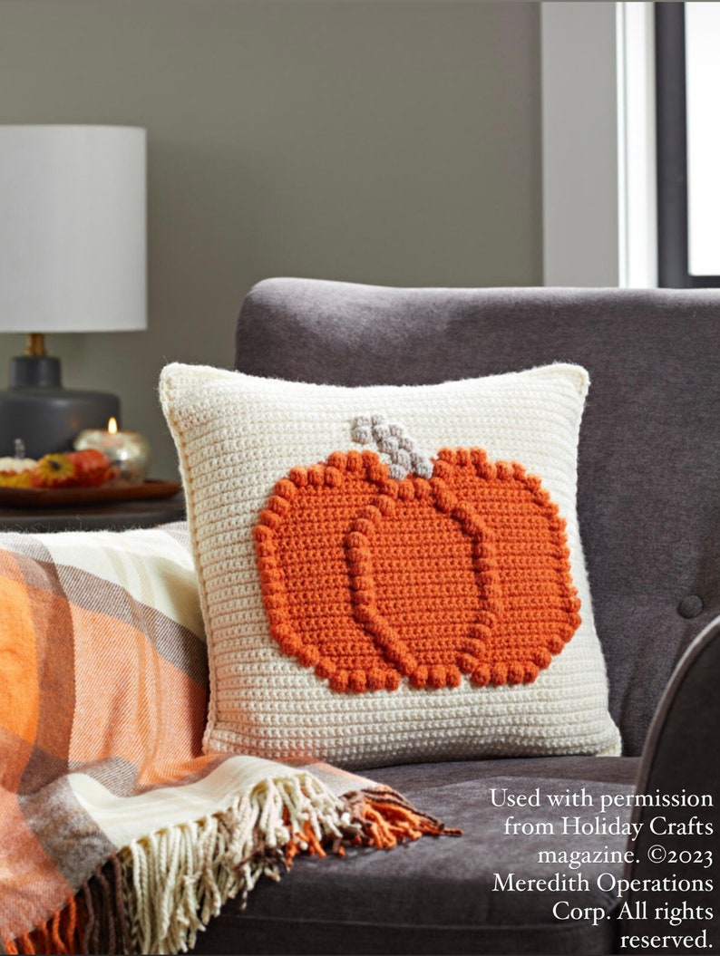 May include: A square, cream-colored crochet pillow featuring a textured, raised orange pumpkin design in the center with a small grey stem. The pumpkin is constructed with a bobble stitch pattern, creating a distinct, three-dimensional appearance.