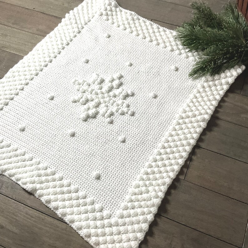 May include: White crocheted throw blanket with a snowflake design. The blanket has a textured, puffy surface.