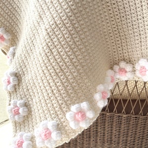 May include: A crocheted baby blanket in cream with a border of white and pink flowers.