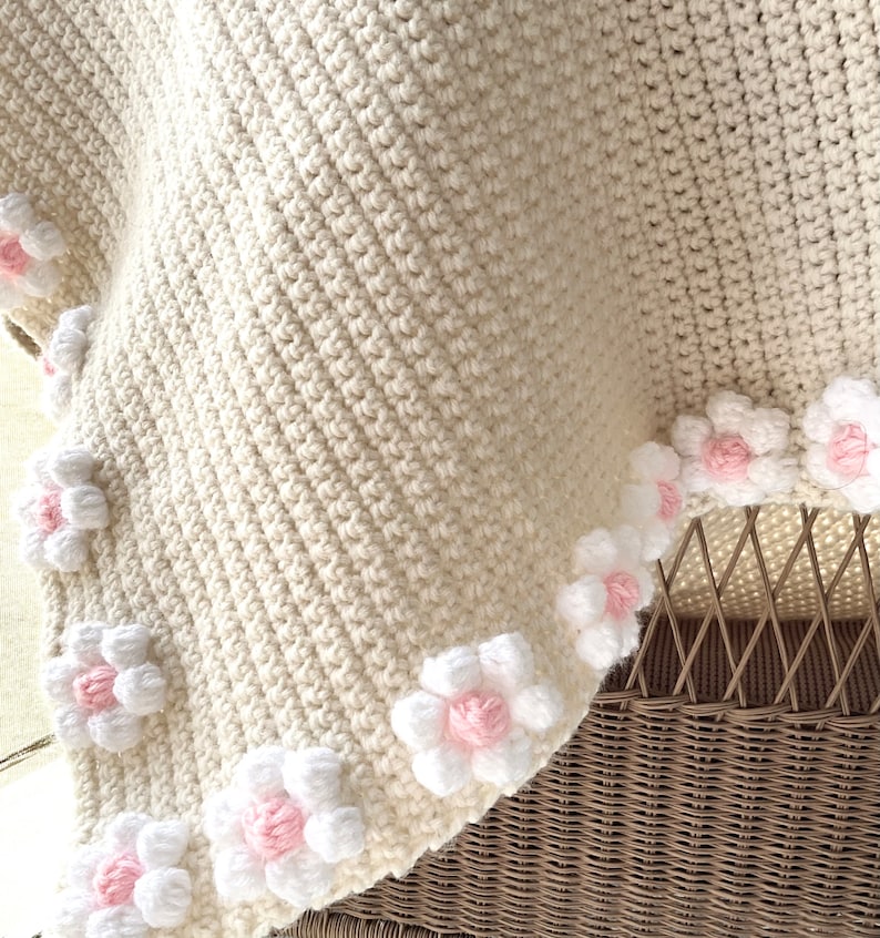 May include: A crocheted baby blanket in cream with a border of white and pink flowers.