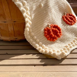 May include: Cream-colored crocheted blanket with orange pumpkin appliqu&eacute;s. The blanket features a textured stitch pattern and a decorative border with bobble details. The pumpkins are made of textured yarn, adding a seasonal touch to the cozy blanket.