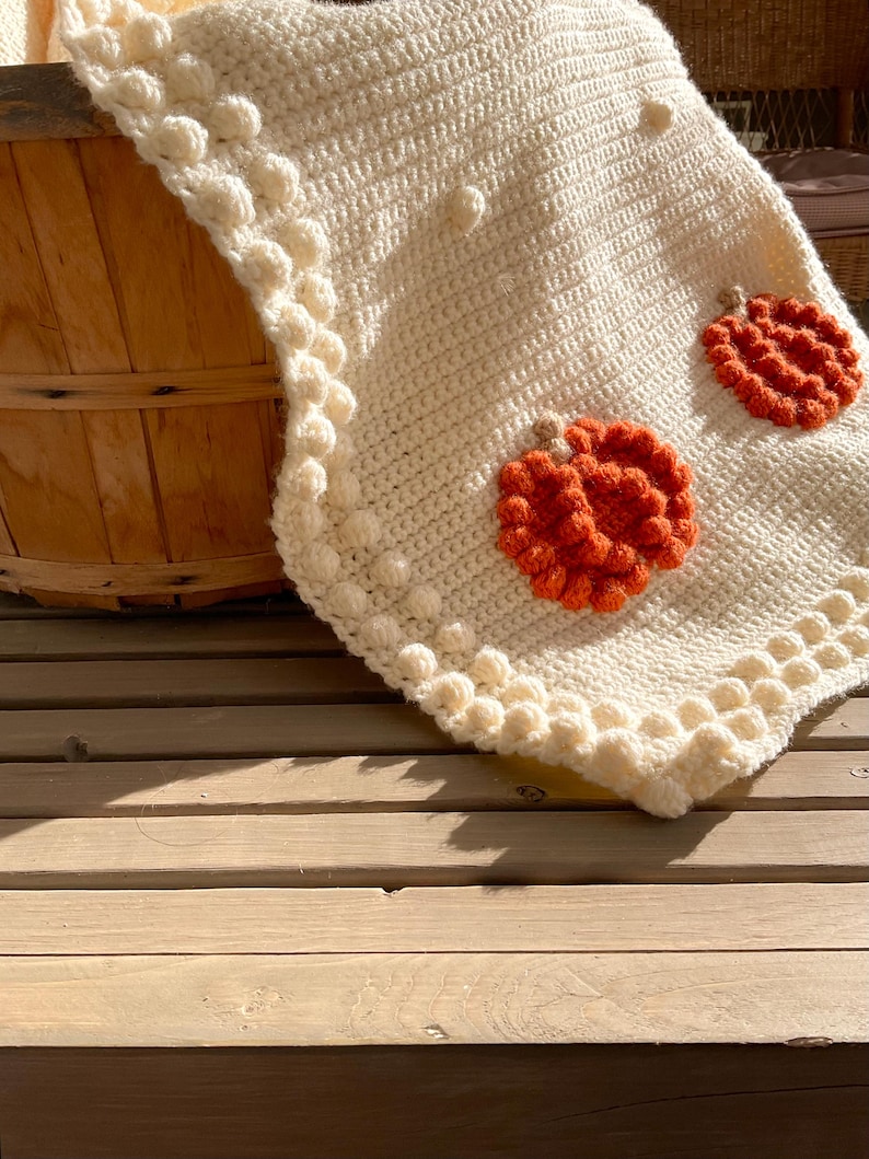 May include: Cream-colored crocheted blanket with orange pumpkin appliqu&eacute;s. The blanket features a textured stitch pattern and a decorative border with bobble details. The pumpkins are made of textured yarn, adding a seasonal touch to the cozy blanket.