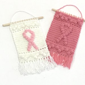 Puede incluir: Dos tapices de pared de ganchillo con listones de madera en la parte superior. El tapiz blanco tiene un dise&ntilde;o de cinta rosa y flecos blancos. El tapiz rosa tiene un patr&oacute;n de coraz&oacute;n y flecos rosas.