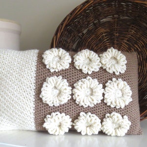 May include: A white and brown crocheted pillow with a decorative pattern of white flowers. The pillow is sitting on a white surface.