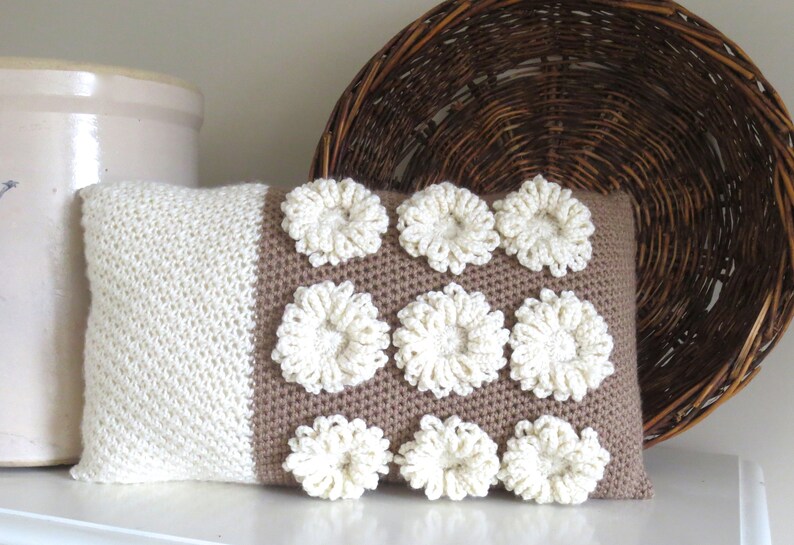 May include: A white and brown crocheted pillow with a decorative pattern of white flowers. The pillow is sitting on a white surface.