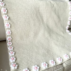 May include: A crocheted baby blanket with a cream background and a border of white and pink flowers.