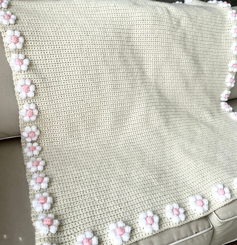 May include: A crocheted baby blanket with a cream background and a border of white and pink flowers.