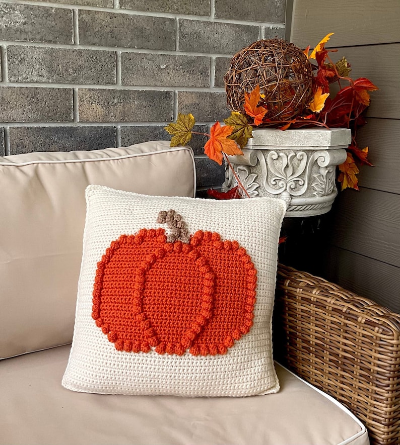 May include: A square cream-colored crochet pillow featuring a large, textured orange pumpkin design with a brown stem centered on the front. The pumpkin is crafted with raised bobble stitches along its edges, creating a distinct, tactile appearance.
