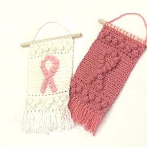 Puede incluir: Dos tapices de pared de ganchillo con listones de madera en la parte superior. Uno es blanco con un dise&ntilde;o de cinta rosa y el otro es rosa con un patr&oacute;n texturizado. Ambos tienen flecos en la parte inferior.