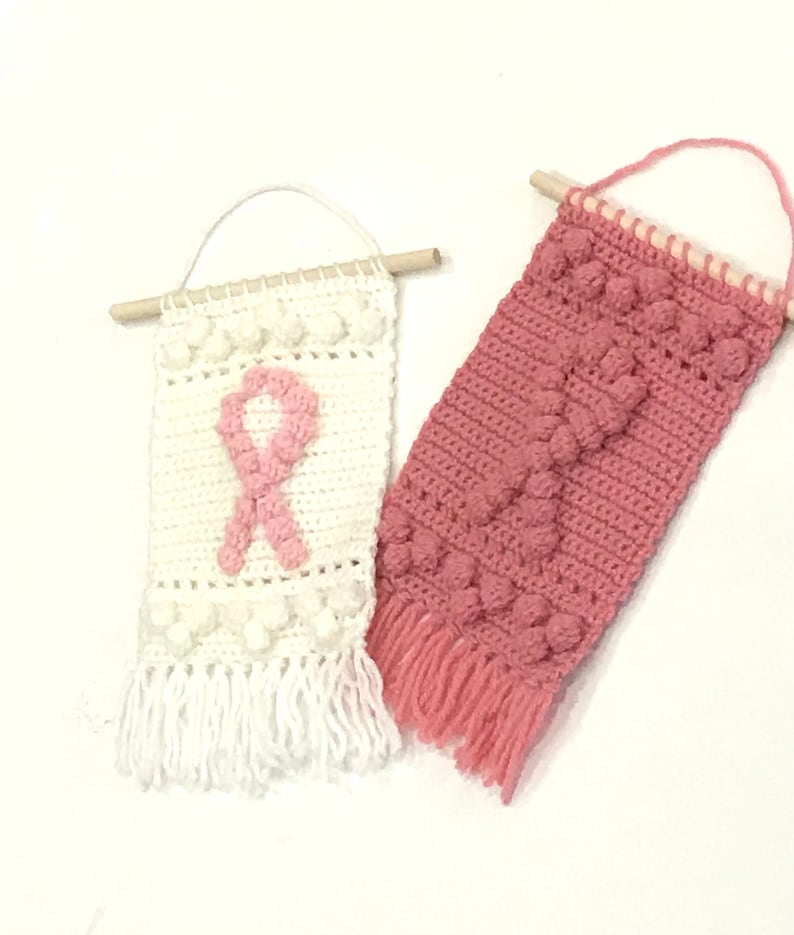Puede incluir: Dos tapices de pared de ganchillo con listones de madera en la parte superior. Uno es blanco con un dise&ntilde;o de cinta rosa y el otro es rosa con un patr&oacute;n texturizado. Ambos tienen flecos en la parte inferior.