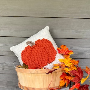 May include: A square white crochet pillow featuring a large, textured orange pumpkin design with a brown stem. The pillow is nestled inside a light-colored wooden basket adorned with artificial autumn leaves in shades of red, orange, and yellow.