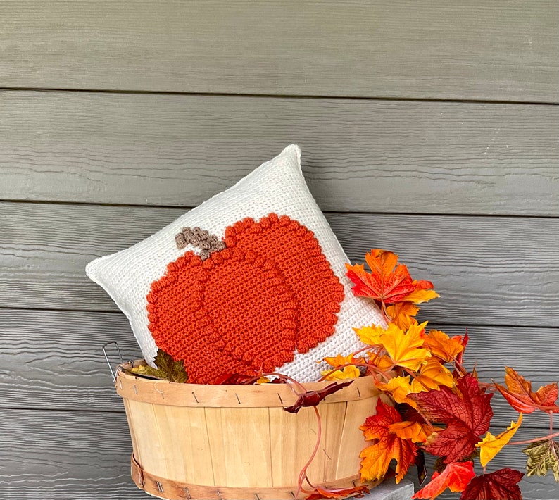 May include: A square white crochet pillow featuring a large, textured orange pumpkin design with a brown stem. The pillow is nestled inside a light-colored wooden basket adorned with artificial autumn leaves in shades of red, orange, and yellow.