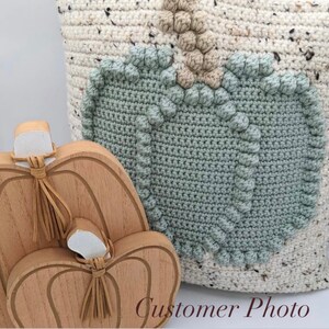 May include: A light-colored speckled crochet bag featuring a large sage green pumpkin motif with a tan stem, positioned next to two stacked wooden pumpkin cutouts with tan leather tassels tied around their stems and white painted tops.