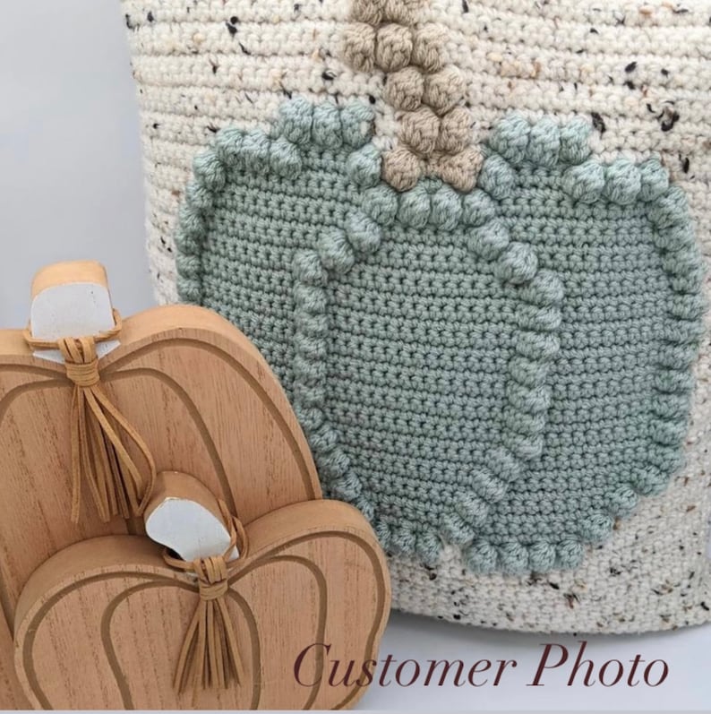 May include: A light-colored speckled crochet bag featuring a large sage green pumpkin motif with a tan stem, positioned next to two stacked wooden pumpkin cutouts with tan leather tassels tied around their stems and white painted tops.