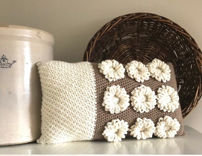 May include: A brown and white crocheted pillow with a textured design. The pillow has a brown background with white crocheted flowers.