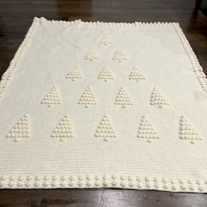 May include: A white crocheted blanket with a design of multiple Christmas trees. The trees are made with raised stitches and are arranged in a pyramid shape.