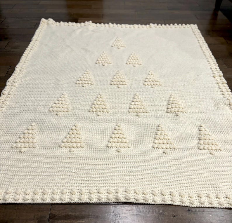 May include: A white crocheted blanket with a design of multiple Christmas trees. The trees are made with raised stitches and are arranged in a pyramid shape.