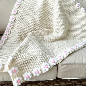May include: A crocheted baby blanket in cream with a border of white and pink flowers.