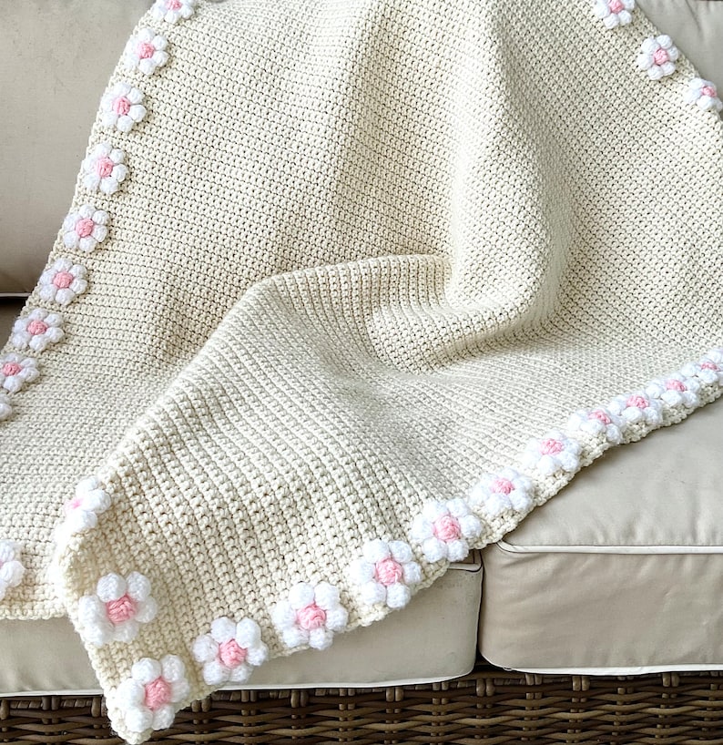 May include: A crocheted baby blanket in cream with a border of white and pink flowers.