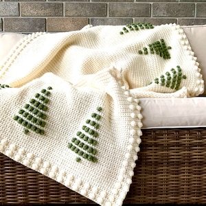 May include: A white crocheted throw blanket with a green and white Christmas tree design. The blanket is draped over a wicker chair.