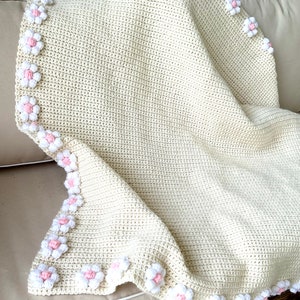 May include: A crocheted baby blanket in cream with a border of pink and white flowers.